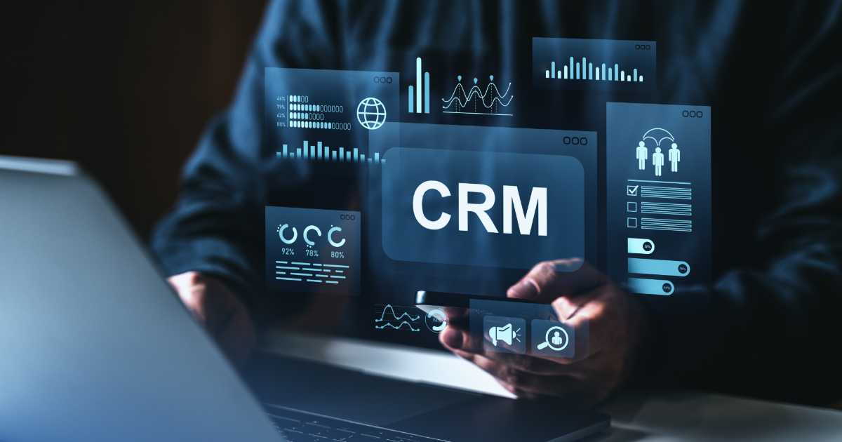 Pessoa usando notebook e smartphone enquanto gráficos digitais e o termo “CRM” aparecem sobrepostos, representando tecnologia e gestão de relacionamento com clientes.