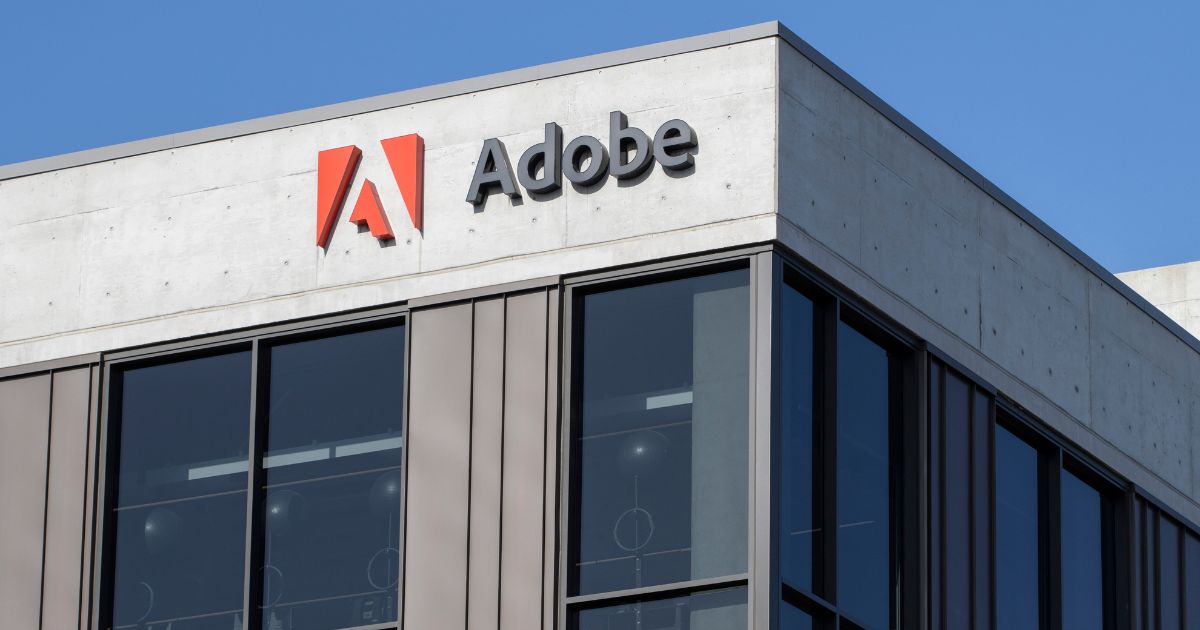 Adobe, marketing, Fachada de um prédio corporativo moderno com o logotipo da Adobe no topo, exibindo o símbolo vermelho e o nome da empresa em letras pretas sobre uma estrutura de concreto claro e janelas amplas de vidro, sob um céu azul