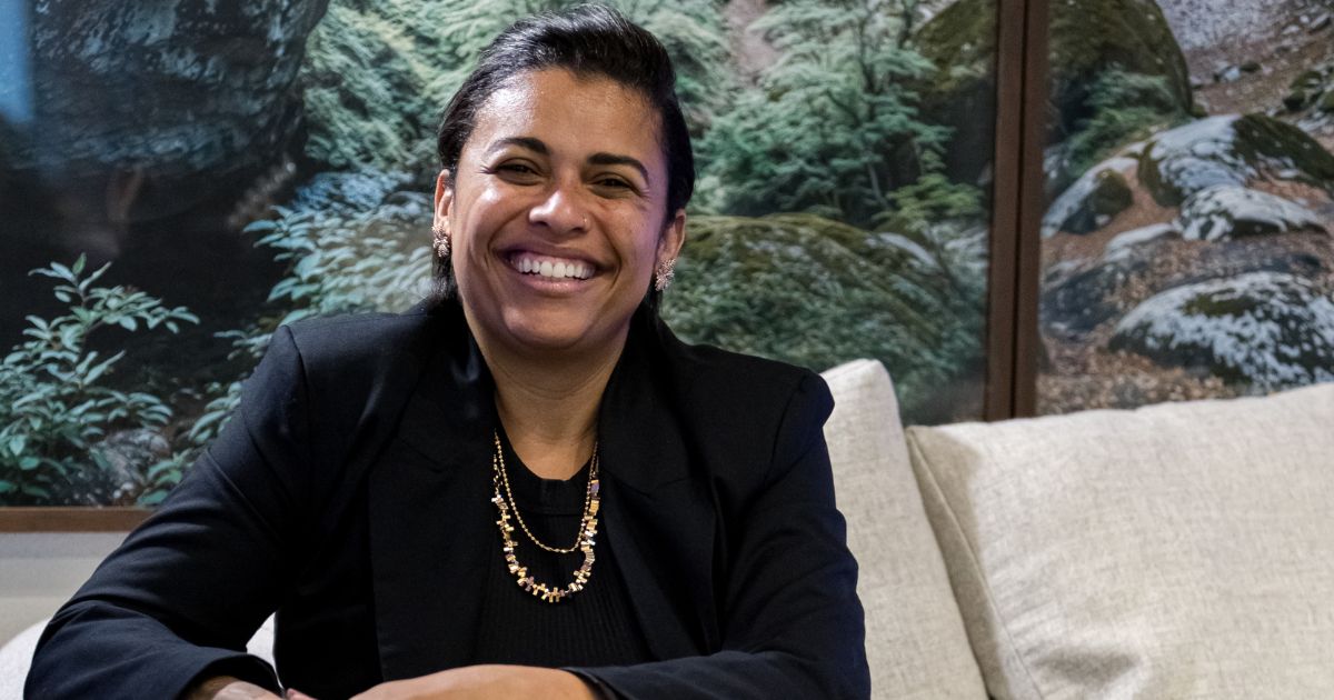 Mulher sorridente sentada em um sofá claro, usando blazer preto e colar dourado, com um painel de paisagem natural ao fundo; junto à imagem aparece o texto “Noéle Gomes, pesquisadora”.
