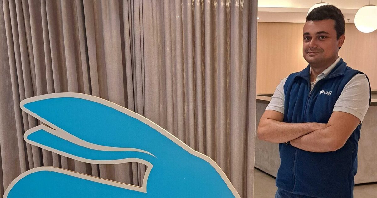 Diego Cançado, da Loggi, está de braços cruzados, vestindo colete azul com o logo da Loggi, posa ao lado de um grande símbolo azul da empresa em um ambiente interno iluminado.