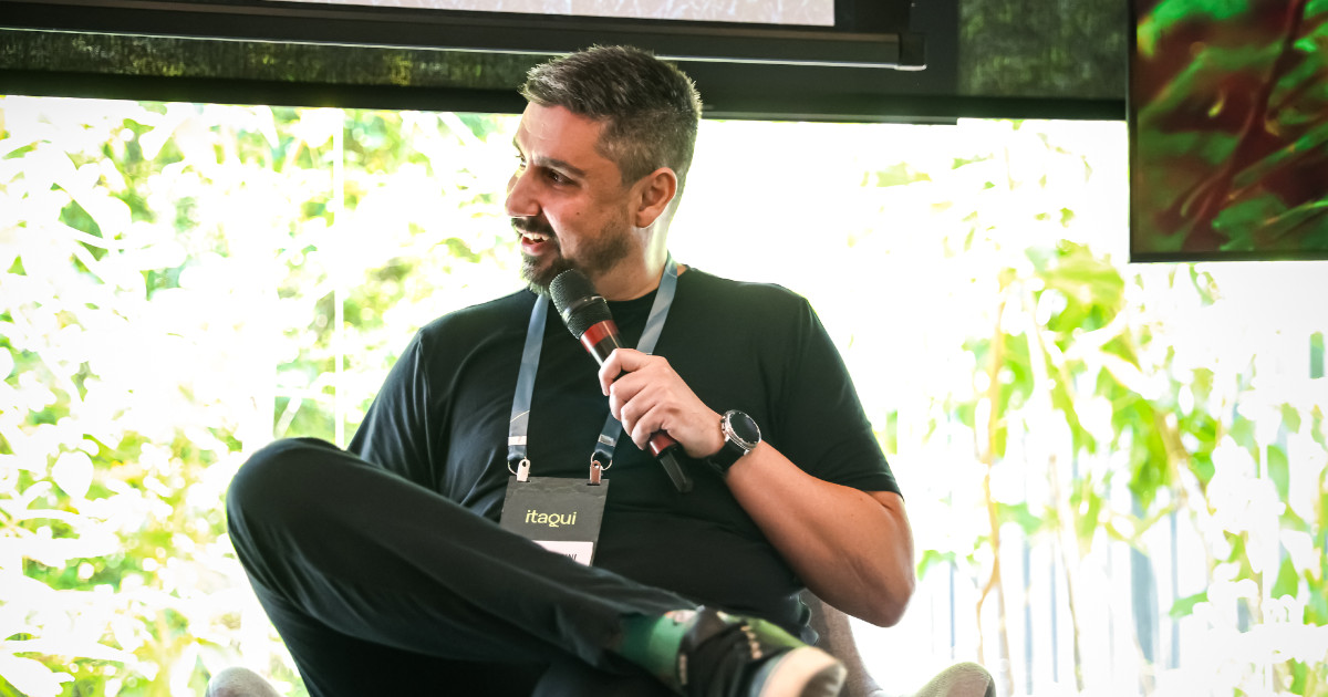 Homem sentado em uma cadeira, segurando um microfone e sorrindo enquanto participa de uma conversa. Ele usa camiseta preta, crachá com o nome “Itaqui” e um relógio de pulso. Ao fundo, há uma janela com vegetação visível. A legenda informa que é Julio Signorini, consultor de cibersegurança do Banco Interamericano de Desenvolvimento (BID). (CISO)