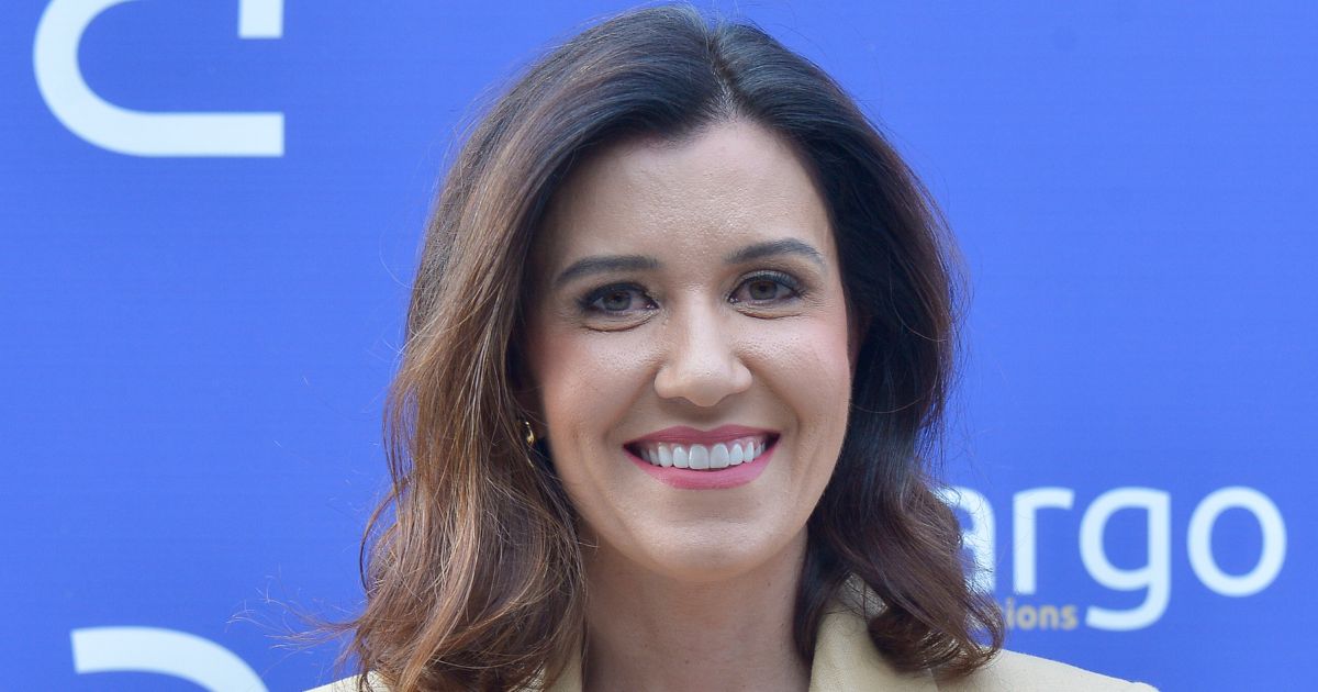 A imagem mostra uma pessoa com cabelos castanhos, ondulados e comprimento médio, vestindo um blazer claro. O fundo é azul, com parte de um logotipo visível à direita, contendo as letras “argo” e um texto menor abaixo, sugerindo que seja um painel ou backdrop de evento corporativo ou conferência.