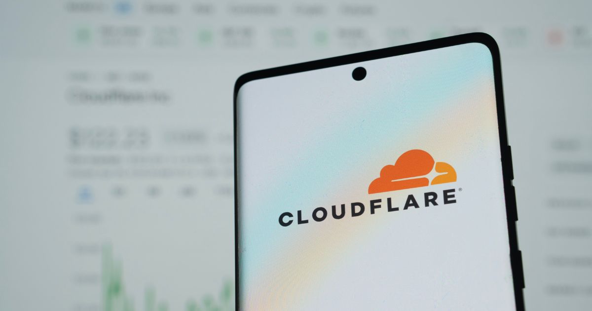 A imagem mostra um smartphone em primeiro plano exibindo o logotipo da Cloudflare, uma empresa conhecida por serviços de segurança, desempenho e confiabilidade na internet. O fundo está desfocado, mas parece mostrar uma tela com gráficos e números, possivelmente relacionados a dados financeiros ou estatísticas.