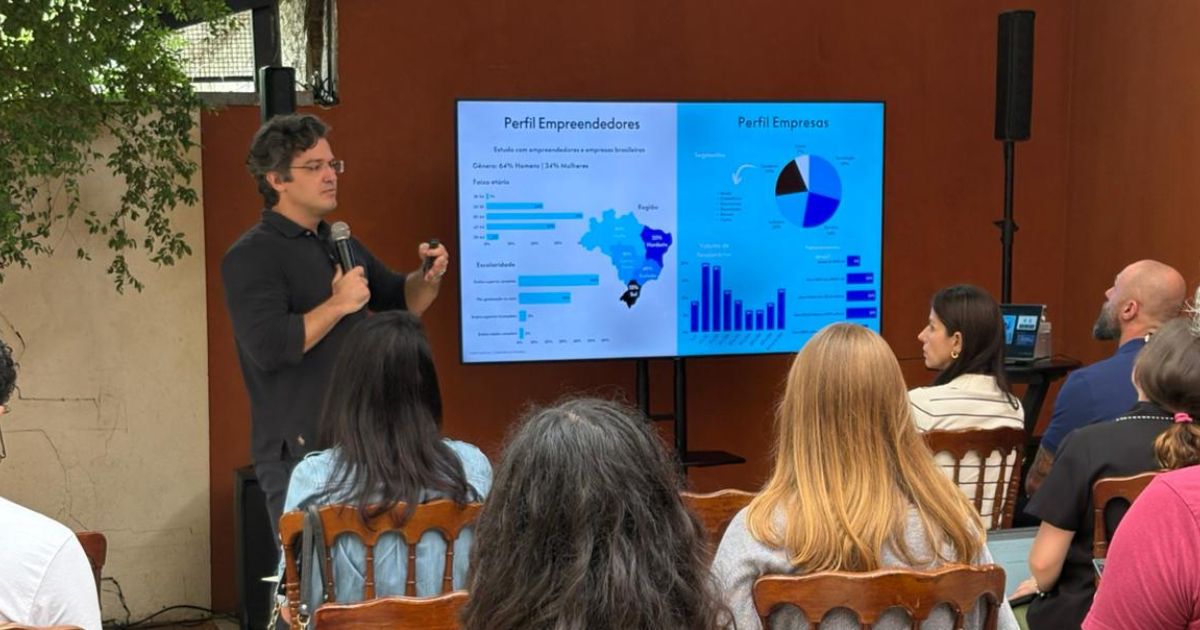 A foto mostra uma apresentação em um espaço aberto com várias pessoas sentadas em cadeiras de madeira, voltadas para uma tela grande. A tela exibe gráficos e textos em português com os títulos “Perfil Empreendedores” e “Perfil Empresas”, incluindo um mapa do Brasil, barras horizontais, um gráfico de pizza e um gráfico de colunas. À esquerda da tela, uma pessoa está em pé segurando um microfone e apontando para os dados exibidos. O ambiente tem paredes em tom terroso e uma árvore parcialmente visível à esquerda, sugerindo um local externo ou semiaberto. (PayPal)