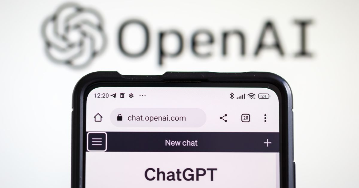 A imagem mostra um smartphone exibindo a página chat.openai.com, com a interface do ChatGPT aberta e o título “New chat” visível na parte superior. O dispositivo está em primeiro plano, com detalhes como ícones de bateria, sinal e hora na barra superior. Ao fundo, desfocado, aparece o logotipo da OpenAI, composto pelo nome “OpenAI” em letras pretas e o símbolo característico da marca. A composição sugere um contexto relacionado a inteligência artificial e tecnologia, destacando a integração entre dispositivos móveis e plataformas de IA.