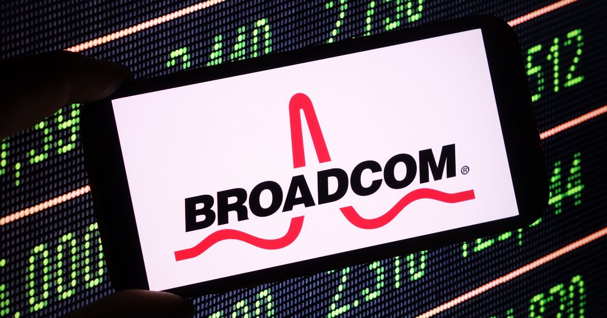 A imagem mostra um smartphone exibindo o logotipo da Broadcom, com o nome da empresa em letras maiúsculas pretas e um símbolo vermelho estilizado acima, sobre um fundo branco. O telefone está inclinado, ocupando a parte central da composição. Ao fundo, há um painel digital com números verdes sobre fundo escuro, lembrando uma tela de cotações ou mercado financeiro, sugerindo um contexto relacionado a tecnologia e investimentos. A iluminação destaca bem o contraste entre o logotipo e os números do painel, reforçando a ideia de negócios e inovação.
