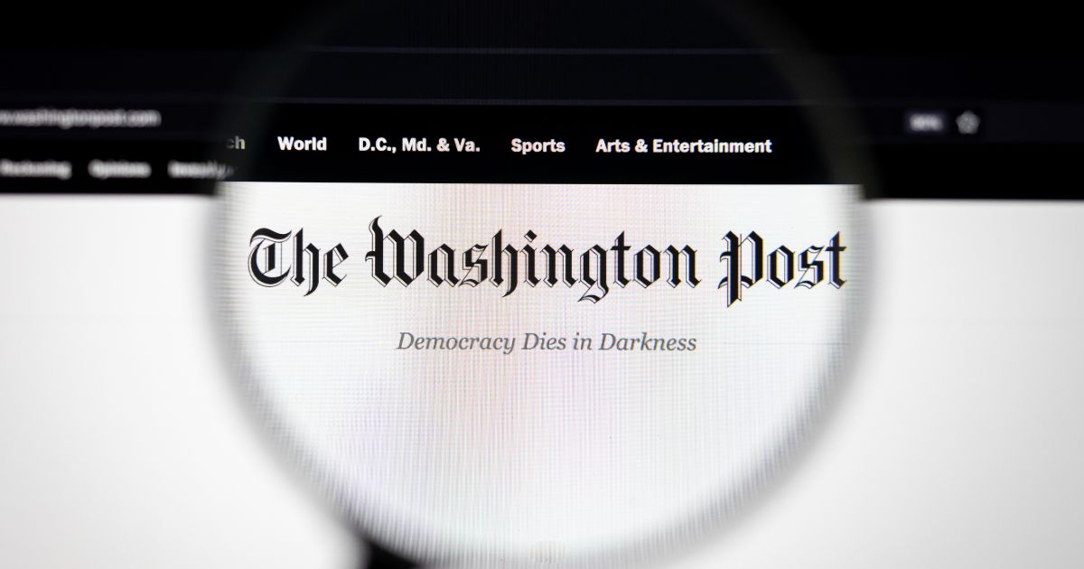 A foto mostra uma tela com o site do jornal The Washington Post ampliado por uma lente de aumento. O logotipo do jornal aparece em destaque na parte superior, com a frase “Democracy Dies in Darkness” logo abaixo. Na barra de navegação superior, são visíveis seções como “World”, “D.C., Md. & Va.”, “Sports” e “Arts & Entertainment”. O fundo é predominantemente branco, com detalhes em preto, e a lente cria um efeito de foco sobre o nome do jornal. (Oracle)