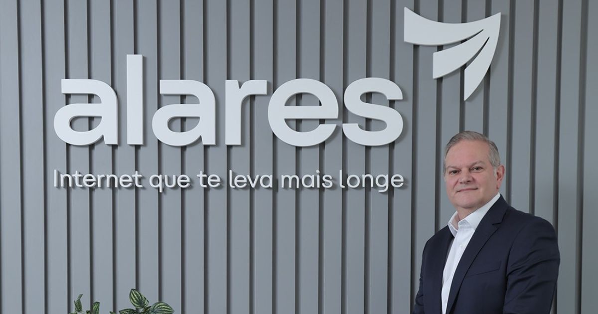 A imagem mostra um ambiente corporativo com uma parede de fundo composta por painéis verticais em tom cinza, onde está fixado o logotipo da empresa “alares” em letras grandes e brancas. Abaixo do nome, aparece o slogan “Internet que te leva mais longe”, também em branco, reforçando a proposta da marca. O design é moderno e minimalista, transmitindo profissionalismo e identidade visual bem definida. A iluminação é suave, destacando o logotipo e o slogan, que são os elementos centrais da composição.