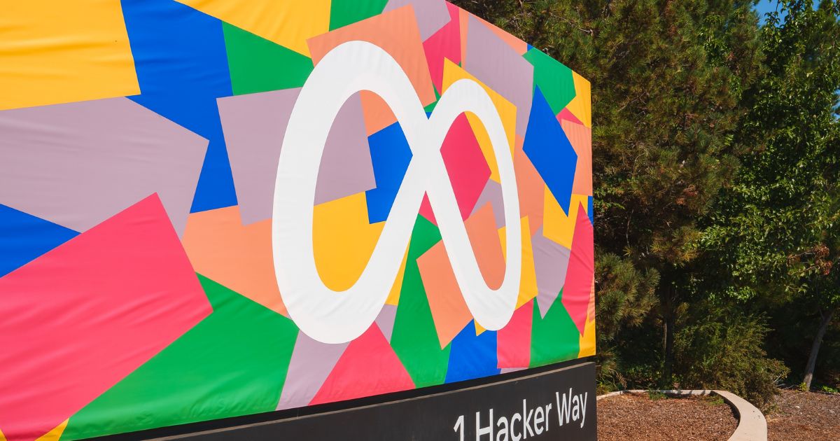 A foto mostra um grande painel colorido com formas geométricas em tons vibrantes, como vermelho, verde, azul, amarelo e rosa. No centro do painel está o logotipo da Meta, representado pelo símbolo de infinito em branco. Na parte inferior do painel, sobre uma base preta, aparece o texto “1 Hacker Way”, indicando o endereço da sede da empresa. Ao fundo, há árvores e vegetação, sugerindo um ambiente externo ensolarado.