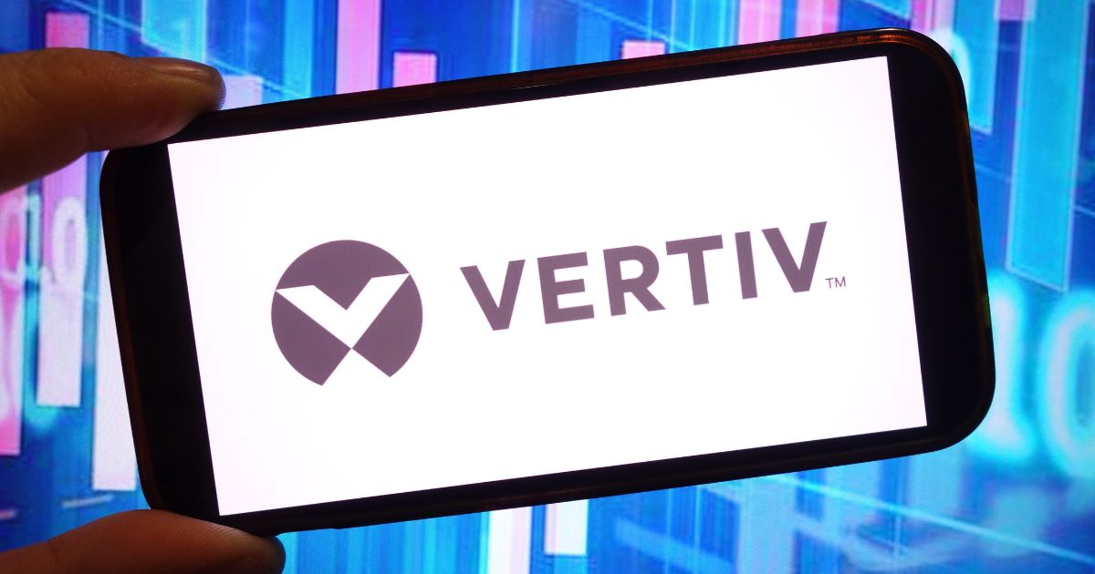 A foto mostra um smartphone sendo segurado na horizontal, exibindo na tela o logotipo da empresa Vertiv. O logotipo consiste em um símbolo geométrico estilizado à esquerda e a palavra “VERTIV” em letras maiúsculas à direita, ambos em cor escura sobre fundo branco. Ao fundo, desfocado, há um cenário com tons de azul e rosa, sugerindo gráficos ou elementos digitais, criando uma atmosfera tecnológica.
