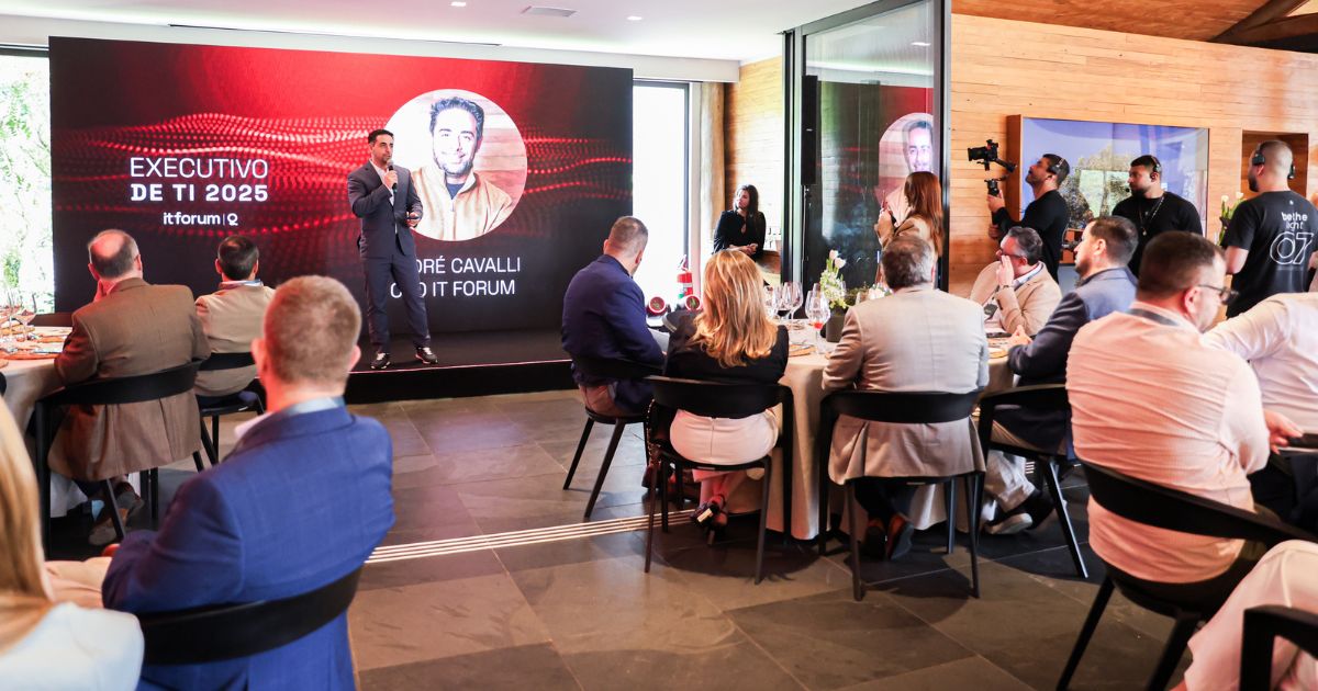 A cena mostra um evento corporativo ou conferência em um espaço fechado, com várias pessoas sentadas ao redor de mesas redondas, acompanhando uma apresentação. No fundo, há um grande painel digital com design em tons de vermelho e preto exibindo o texto: “EXECUTIVO DE TI 2025 itforumIQ ANDRÉ CAVALLI CEO IT FORUM” À frente do painel, uma pessoa está em pé, segurando um microfone, aparentemente fazendo um discurso ou apresentação. À direita, há uma equipe de apoio, incluindo um fotógrafo e outras pessoas em pé, observando a cena. As mesas estão organizadas com toalhas claras, arranjos de flores e copos, sugerindo um ambiente formal e elegante. A iluminação é natural, vinda de grandes janelas à esquerda, complementada por luz artificial. (vencedores)