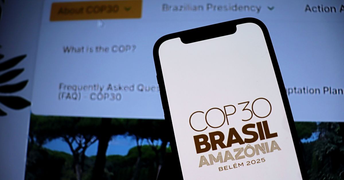 A cena mostra um smartphone em primeiro plano exibindo o logotipo e texto “COP30 BRASIL AMAZÔNIA BELÉM 2025” em letras marrons sobre fundo claro. Ao fundo, desfocado, há uma tela com informações sobre a COP30, incluindo seções como “About COP30”, “Brazilian Presidency”, e perguntas frequentes (FAQ – COP30). A composição sugere um contexto relacionado à conferência climática COP30, destacando sua realização no Brasil, na Amazônia, em Belém, no ano de 2025.