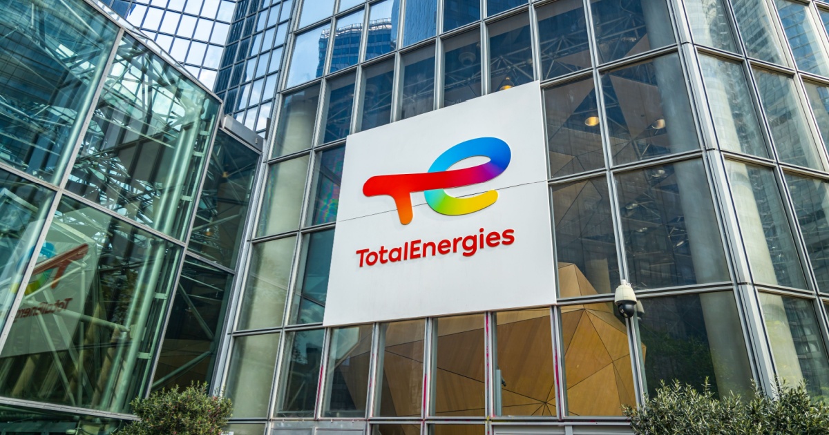 Fachada de um prédio de vidro moderno com o logotipo da empresa TotalEnergies exibido em um grande painel branco, mostrando as letras “TE” em cores vibrantes e o nome “TotalEnergies” em vermelho abaixo.