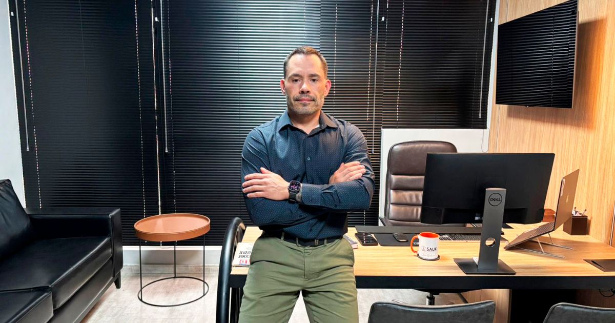 Um homem está em pé, de braços cruzados, em um escritório moderno. Ele usa camisa social azul escura e calça verde. Atrás dele há uma mesa de madeira clara com um monitor, teclado, mouse, caneca, smartphone e outros itens organizados. À esquerda, vê-se um sofá preto e uma pequena mesa redonda. As janelas possuem persianas escuras fechadas e, à direita, há uma TV ou tela fixa na parede de madeira. (TI)