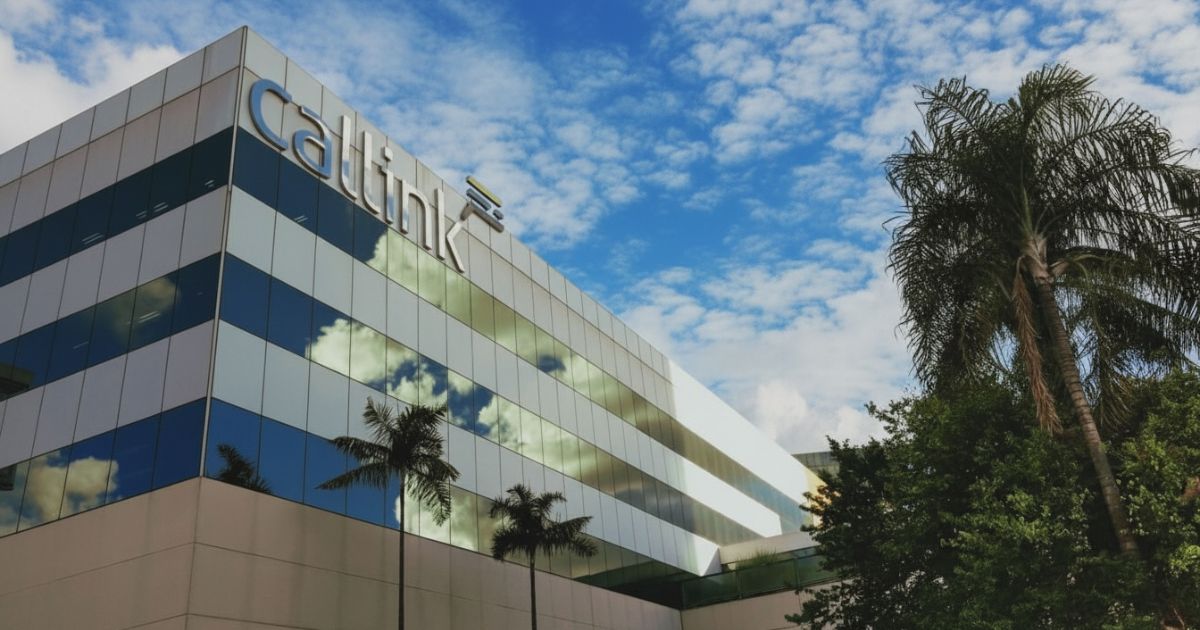 A imagem mostra a fachada de um edifício corporativo moderno com revestimento espelhado, refletindo o céu azul com nuvens brancas. Na parte superior do prédio, está visível o logotipo “Callink” em letras grandes e metálicas. Em frente ao edifício, há palmeiras e árvores, adicionando um toque natural ao cenário. A composição transmite um ambiente empresarial sofisticado, com contraste entre a arquitetura contemporânea e a vegetação.