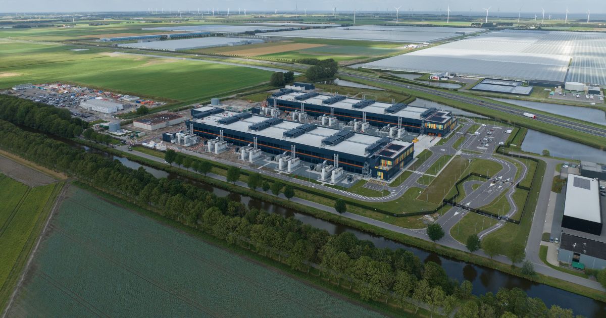 A imagem mostra uma vista aérea de um grande complexo industrial ou centro de dados, localizado em uma área rural. O complexo é composto por vários edifícios retangulares com telhados claros e estruturas externas que parecem sistemas de ventilação ou resfriamento. Ao redor do complexo, há ruas internas pavimentadas, áreas de estacionamento e canais de água que delimitam o terreno. O cenário ao fundo é formado por extensos campos verdes, estufas agrícolas e fileiras de turbinas eólicas visíveis no horizonte, indicando uma região com forte presença de agricultura e geração de energia renovável. O céu está parcialmente nublado, com nuvens brancas dispersas, e a iluminação sugere um dia claro. A imagem transmite uma sensação de infraestrutura moderna integrada a um ambiente rural. (TIP Brasil)