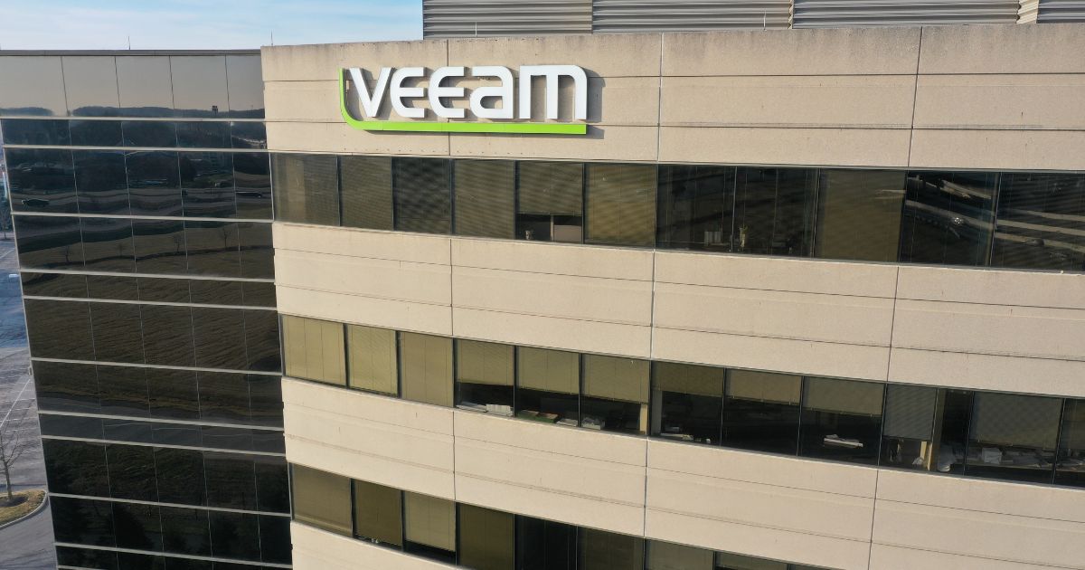 Um prédio comercial moderno aparece em destaque, com fachada composta por painéis de vidro escuro e áreas de concreto claro. Na parte superior da construção, há um letreiro grande com o nome “Veeam” em letras brancas e um detalhe verde sob a marca. As janelas refletem o ambiente externo e revelam parte do interior dos andares. A cena foi capturada de um ângulo levemente elevado, mostrando boa parte da lateral do edifício.