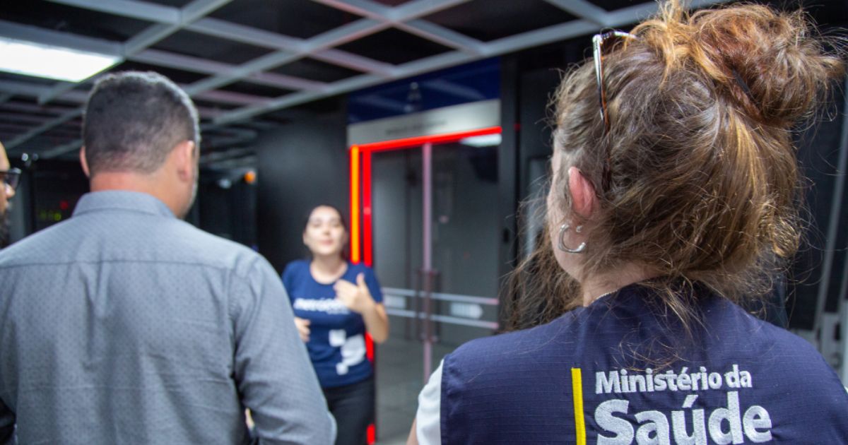 A imagem mostra um ambiente interno com estrutura moderna, teto com painéis quadriculados e iluminação artificial. No centro, há uma área delimitada por um portal com luzes vermelhas, que parece ser um equipamento de controle ou segurança, possivelmente um detector ou scanner. Em primeiro plano, uma pessoa está de costas usando um colete azul com a inscrição “Ministério da Saúde” em letras brancas e amarelas, indicando vínculo com o órgão público brasileiro. Outra pessoa também aparece parcialmente de costas, vestindo uma camisa cinza. Ao fundo, há uma terceira pessoa gesticulando, sugerindo que está explicando ou orientando algo. O cenário transmite a ideia de inspeção, monitoramento ou visita técnica em uma instalação controlada.