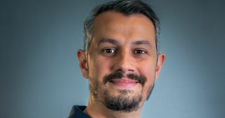 A imagem mostra a parte superior de uma pessoa, enquadrada dos ombros para cima, posicionada diante de um fundo azul acinzentado uniforme. A pessoa veste uma camisa escura com gola simples, e o cabelo curto aparece no topo da imagem. A iluminação é suave e destaca o contraste entre a roupa e o fundo. (André Luiz Lopes Petenussi, CTO da Localiza&co)