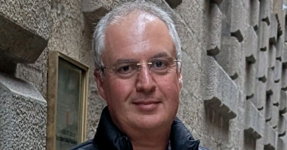 MediaTek, IA A imagem mostra uma pessoa vestindo uma jaqueta preta, posicionada em frente a uma parede de pedra com textura rugosa e blocos salientes, sugerindo um estilo arquitetônico robusto. À esquerda, há uma placa retangular emoldurada, aparentemente metálica, fixada na parede, com alguns elementos gráficos e texto parcialmente visível. O cenário parece ser externo, com iluminação natural, e transmite a ideia de um ambiente urbano ou histórico.