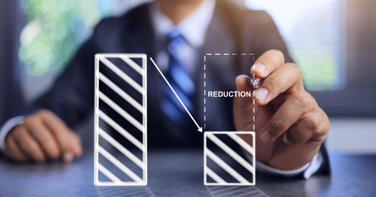 A imagem mostra uma pessoa vestindo um terno escuro com camisa clara e gravata listrada, segurando uma caneta ou marcador. Em primeiro plano, há uma ilustração gráfica composta por duas barras: uma maior à esquerda e outra menor à direita, ambas com preenchimento listrado. Uma seta aponta da barra maior para a menor, indicando uma redução. Ao lado da barra menor, está escrito “REDUCTION”, reforçando a ideia de diminuição ou corte. O fundo é desfocado, destacando a mão e o gráfico como elementos principais, sugerindo um contexto corporativo ou financeiro, relacionado a redução de custos, despesas ou indicadores. (demissões)