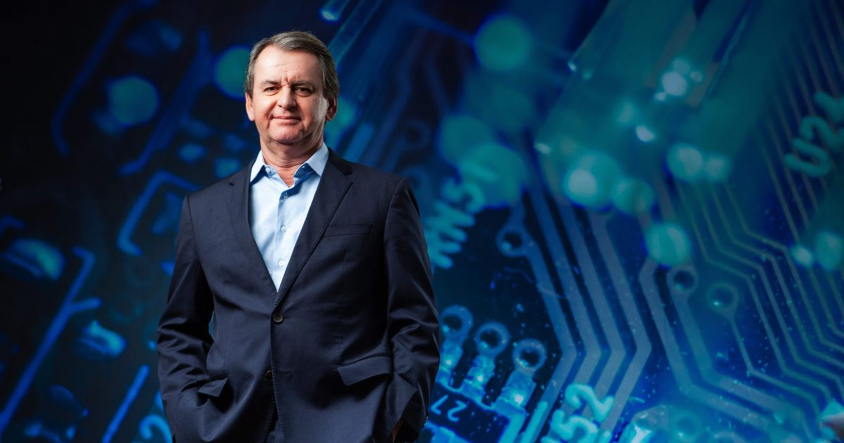 Um homem veste um terno azul-escuro com camisa social clara e está posicionado diante de um grande painel digital que exibe um fundo abstrato inspirado em circuitos eletrônicos, com tons de azul e detalhes luminosos. Ele mantém as mãos nos bolsos do paletó e sua postura transmite confiança, compondo uma cena que remete a tecnologia, inovação e ambiente corporativo. (Ricardo Bloj, presidente da Lenovo Brasil )