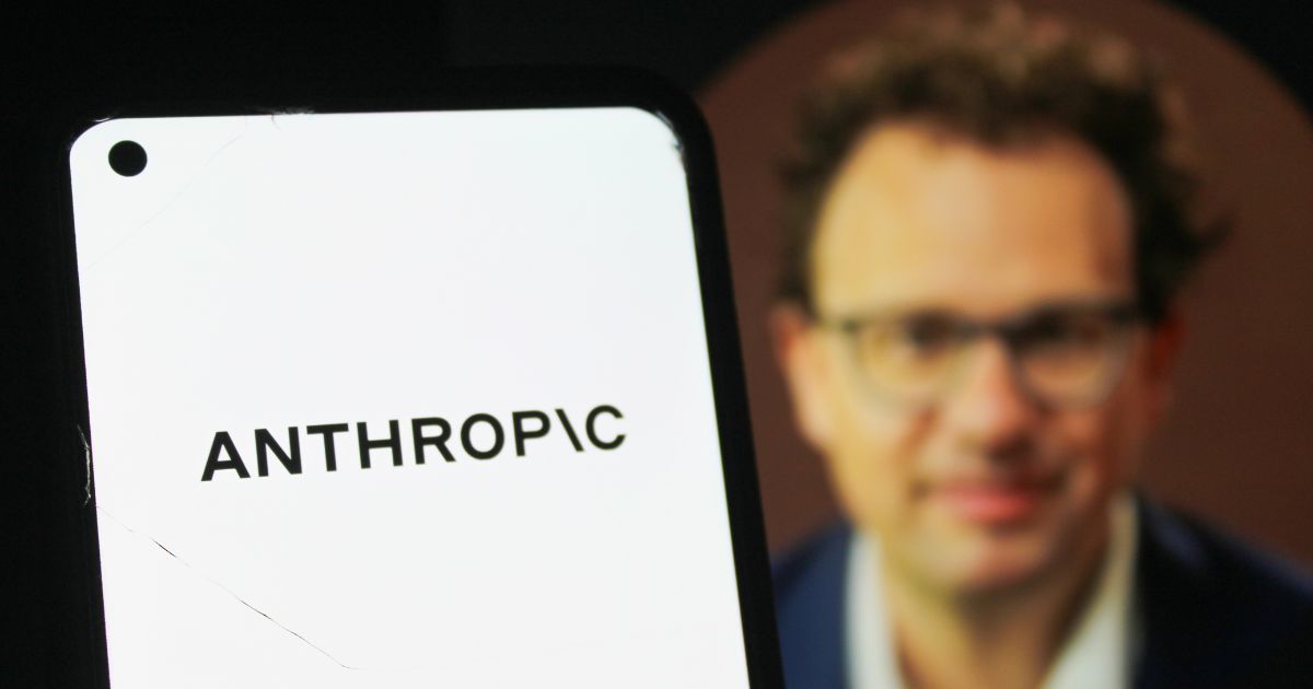 Telefone celular exibindo, em destaque, o nome “ANTHROPIC” em letras pretas sobre fundo branco. Ao fundo, aparece uma pessoa usando roupas formais, em segundo plano, com iluminação suave e foco concentrado no dispositivo móvel. (humanidade)