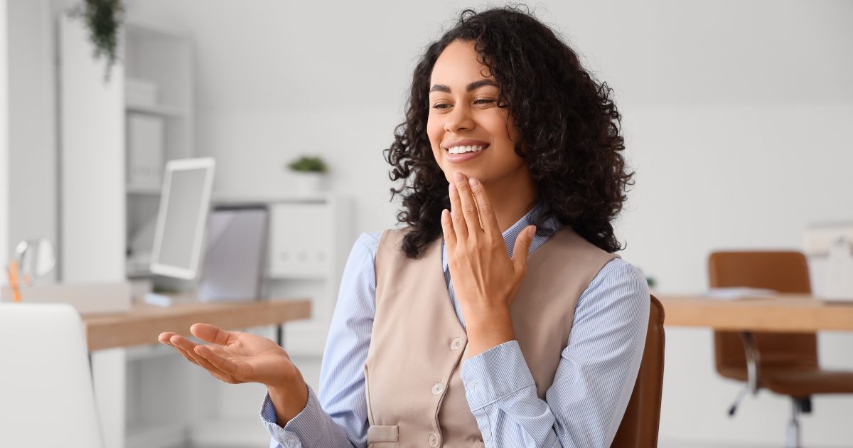 Pessoa sentada em um escritório moderno, vestindo camisa social azul-clara e colete bege. Ela faz um gesto com uma das mãos levantadas e a outra posicionada próximo ao rosto, como se explicasse ou apresentasse algo. Ao fundo, há mesas, computador e cadeiras em tons neutros, compondo um ambiente profissional e organizado. (surda)