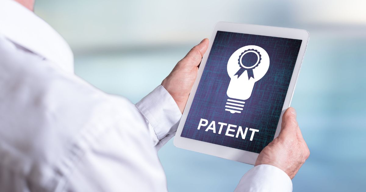 Pessoa segurando um tablet na horizontal, que exibe a imagem de uma lâmpada estilizada com um selo de certificação no centro e a palavra “PATENTE” em destaque abaixo. O fundo do dispositivo tem textura azul escura com linhas sutis, enquanto o ambiente ao redor é claro e desfocado, sugerindo um escritório ou espaço corporativo.