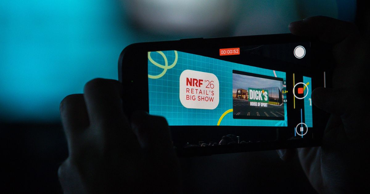 A imagem mostra um close-up de um smartphone sendo usado para gravar ou transmitir um evento. Aqui estão os detalhes: Contexto geral: O telefone está em modo de gravação, exibindo uma tela com elementos gráficos e texto relacionados a um evento. Texto visível na tela do telefone: NRF '26 Retail's Big Show: Indica que se trata do evento NRF 2026, um dos maiores encontros globais sobre varejo. Dick's House of Sport: Provavelmente uma apresentação ou case exibido durante o evento. Elementos visuais: Fundo azul com padrões geométricos e linhas curvas, sugerindo um design moderno e tecnológico. Há também indicadores de gravação (ícone vermelho e cronômetro mostrando 00:00:52). Iluminação e cores: Predominância de tons azulados e pretos, com contraste forte entre a tela do telefone e o ambiente ao redor.