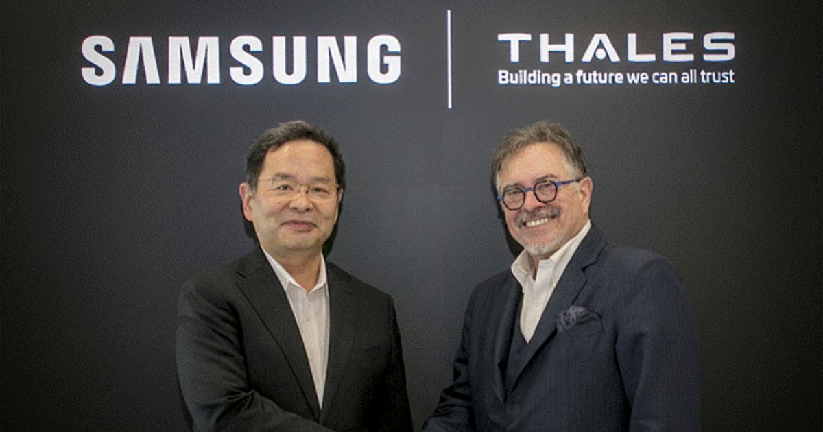 Duas pessoas usando trajes formais apertam as mãos diante de um painel preto com os logotipos “Samsung” e “Thales” acompanhados do slogan “Building a future we can all trust”. A cena representa uma parceria corporativa ou assinatura de acordo entre as duas empresas.