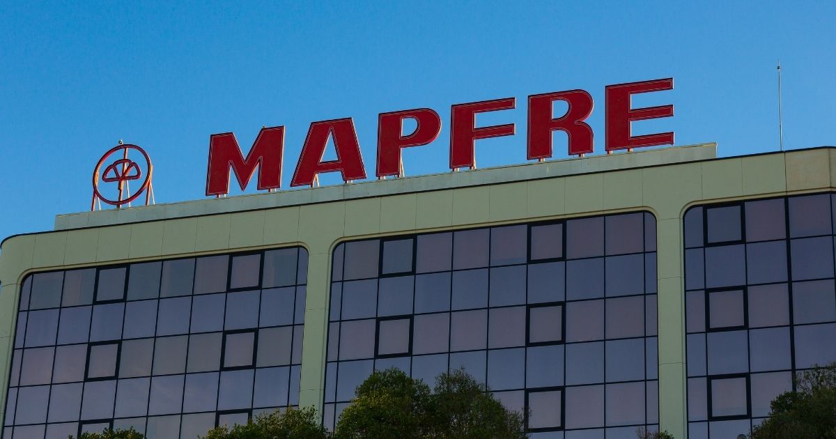 A imagem mostra a fachada de um edifício corporativo com o logotipo e nome MAPFRE em destaque na parte superior. Detalhes principais: Texto visível: “MAPFRE” em letras grandes e vermelhas, acompanhado do símbolo da empresa à esquerda. Arquitetura: Estrutura moderna com linhas retas e revestimento claro. Fileiras de janelas espelhadas, refletindo o céu e parte da vegetação ao redor. Ambiente: Céu limpo e azul, indicando um dia ensolarado. Vegetação na base da imagem, sugerindo árvores próximas ao prédio. Composição visual: O contraste entre o vermelho do logotipo e o azul do céu cria uma imagem marcante e profissional. Essa imagem remete a instituições financeiras, seguros e soluções corporativas, transmitindo solidez e credibilidade.