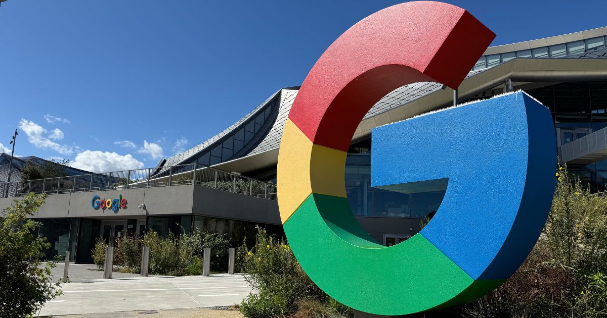 A imagem mostra a entrada de um prédio corporativo com um design moderno e sofisticado, identificado como parte do campus do Google. Detalhes principais: Elemento em destaque: Um grande monumento com a letra “G” estilizada nas cores características do Google (vermelho, azul, verde e amarelo), localizado em frente ao edifício. Arquitetura: Estrutura contemporânea com linhas curvas, amplas janelas de vidro e telhado inclinado, transmitindo inovação e sustentabilidade. Ambiente externo: Céu limpo e azul, vegetação bem cuidada ao redor da entrada, criando um espaço agradável e acolhedor. Texto visível: A palavra “Google” aparece na fachada do prédio, reforçando a identidade da empresa.