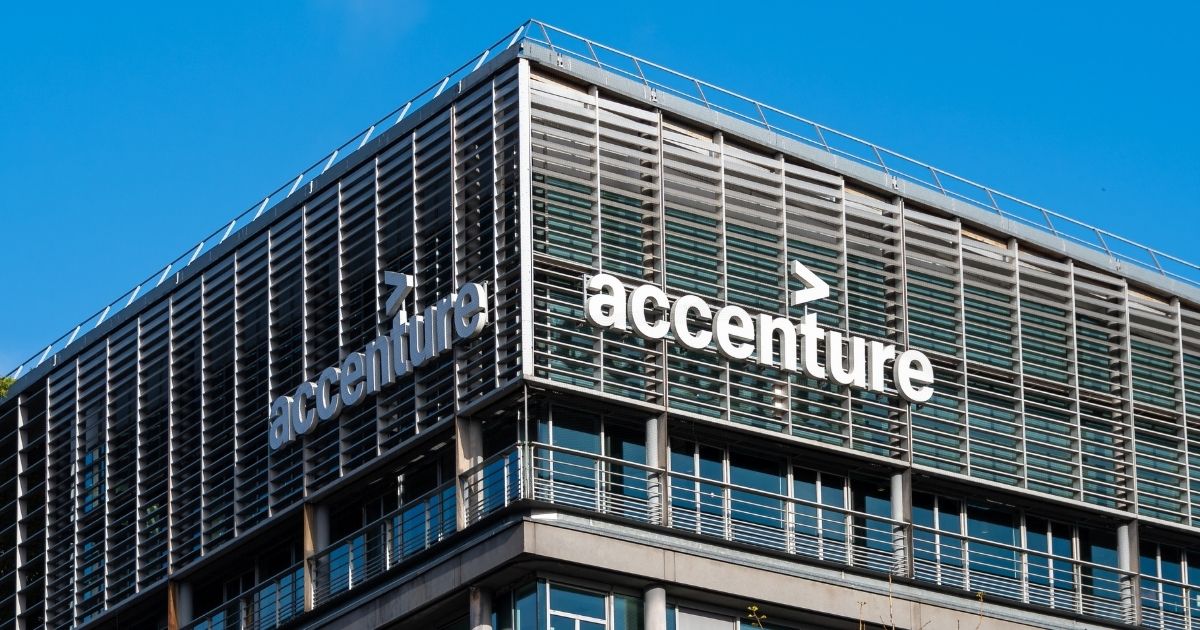 A imagem mostra a fachada de um edifício corporativo com o logotipo da Accenture em destaque. Detalhes principais: Texto visível: “accenture” escrito em letras minúsculas brancas, acompanhado do símbolo característico da empresa (um sinal de maior “>” acima da letra “t”). Arquitetura: Estrutura moderna com painéis horizontais metálicos e vidros, criando um design sofisticado e funcional. Linhas retas e simétricas, reforçando um estilo corporativo contemporâneo. Ambiente: Céu azul claro ao fundo, indicando um dia ensolarado. A imagem transmite uma sensação de profissionalismo e inovação.