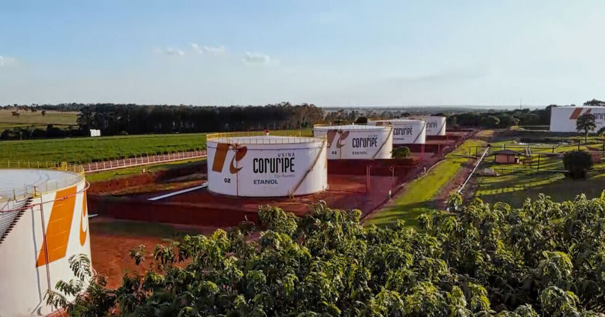 A imagem mostra uma área industrial ao ar livre, composta por diversos tanques cilíndricos de grande porte utilizados para armazenamento. Cada tanque é pintado de branco com detalhes em laranja e exibe a marca “Usina Coruripe” e a palavra “Etanol”. Os tanques estão distribuídos ao longo de um terreno amplo, com solo avermelhado e caminhos internos organizados. Ao redor da instalação, há extensas áreas verdes, incluindo plantações e áreas de vegetação baixa. No horizonte, é possível ver faixas de árvores e o céu claro de fim de tarde, iluminando toda a paisagem com luz natural suave.