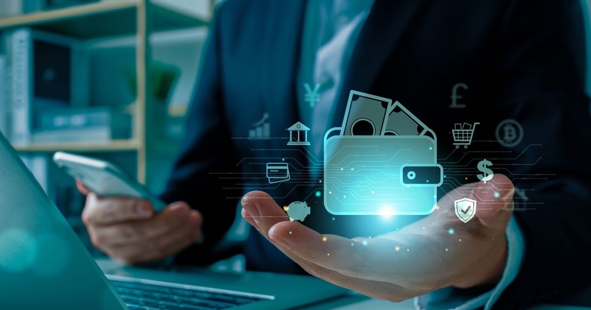 A imagem apresenta um conceito visual relacionado a finanças digitais e tecnologia. Detalhes principais: Elemento central: Uma mão aberta com uma sobreposição digital mostrando um ícone de carteira eletrônica contendo notas de dinheiro. Ao redor da carteira, há ícones que representam diferentes aspectos financeiros, como: Símbolos monetários (dólar, libra, iene, bitcoin). Ícones de segurança (escudo com check). Carrinho de compras (e-commerce). Gráficos e indicadores financeiros. Contexto físico: A pessoa segura um smartphone na outra mão, sugerindo uso de aplicativos financeiros ou pagamentos digitais. Um laptop está parcialmente visível sobre a mesa, reforçando a ideia de integração tecnológica. Traje formal (terno), indicando um ambiente corporativo ou profissional. Cores e iluminação: Tons escuros no cenário físico contrastando com os elementos digitais em azul neon, criando uma estética futurista e tecnológica. Essa imagem transmite inovação em serviços financeiros, transformação digital, segurança e praticidade nos pagamentos online. (crédito)