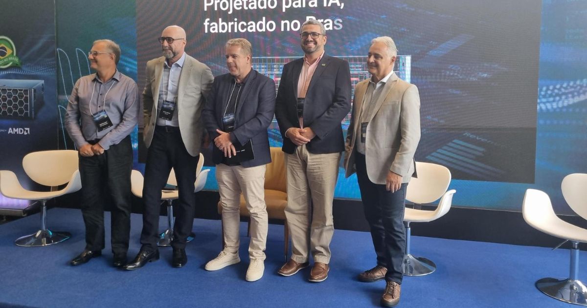 Cinco pessoas em pé, lado a lado, sobre um palco de evento corporativo. Elas usam roupas sociais e crachás de identificação. Atrás delas há um grande painel digital com textos e gráficos tecnológicos. Cadeiras brancas de design moderno estão dispostas ao fundo, sugerindo um painel ou apresentação oficial. (Dell Technologies)