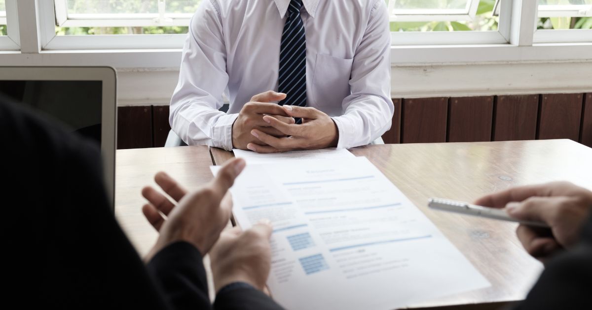 A imagem mostra uma cena típica de entrevista ou reunião profissional em um ambiente corporativo. Detalhes principais: Primeiro plano: Duas mãos segurando um currículo ou documento impresso, com texto visível em formato de lista, indicando que é provavelmente um CV ou proposta. Outra mão segura uma caneta, sugerindo anotações ou avaliação. Centro da imagem: Pessoa vestindo camisa social branca e gravata listrada azul, com as mãos entrelaçadas sobre a mesa, postura formal e atenta. Ambiente: Mesa de madeira clara, com um laptop parcialmente visível à esquerda. Fundo com janelas amplas, permitindo entrada de luz natural, e plantas do lado externo, criando um clima profissional e acolhedor. Essa imagem transmite claramente processos seletivos, entrevistas de emprego ou reuniões de negócios.