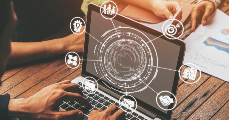 Pessoa digitando em um notebook apoiado sobre uma mesa de madeira. Na tela, uma interface gráfica em estilo holográfico exibe um globo central conectado a ícones circulares que representam colaboração, estratégia, inovação, metas, trabalho em equipe e tomada de decisões. Ao redor, há documentos com gráficos e relatórios, sugerindo análise de dados e planejamento estratégico em um contexto corporativo e tecnológico. (transformação digital)