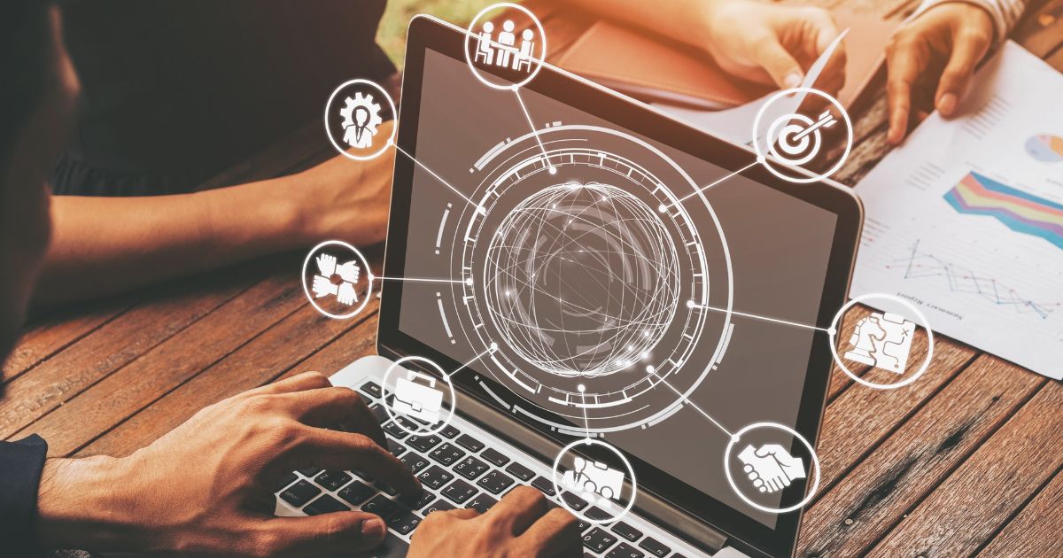 Pessoa digitando em um notebook apoiado sobre uma mesa de madeira. Na tela, uma interface gráfica em estilo holográfico exibe um globo central conectado a ícones circulares que representam colaboração, estratégia, inovação, metas, trabalho em equipe e tomada de decisões. Ao redor, há documentos com gráficos e relatórios, sugerindo análise de dados e planejamento estratégico em um contexto corporativo e tecnológico. (transformação digital)
