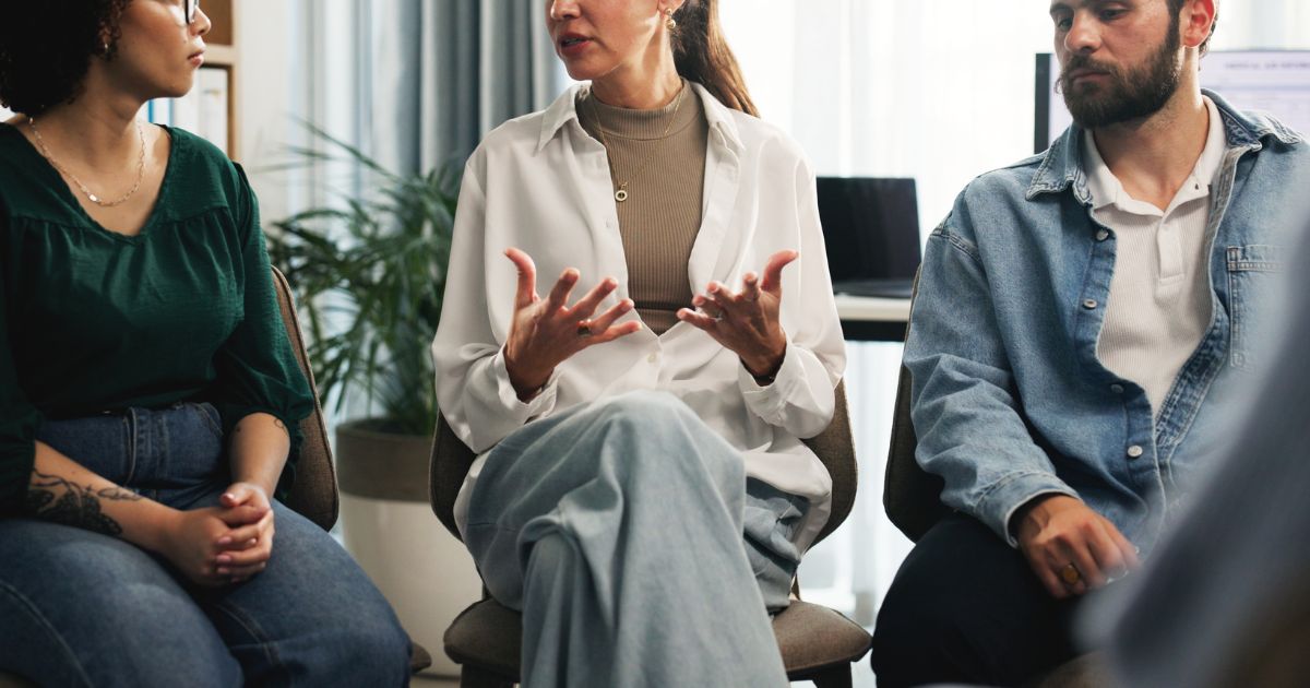 Três pessoas sentadas em cadeiras, participando de uma conversa em grupo. A pessoa ao centro gesticula com as mãos enquanto fala, usando camisa clara e calça ampla. À esquerda, uma pessoa com blusa verde e jeans escuros escuta atentamente; à direita, outra pessoa veste jaqueta jeans clara sobre camisa branca. Ao fundo, há plantas e cortinas, sugerindo um ambiente acolhedor e de diálogo, como uma sessão de apoio ou reunião colaborativa. (janeiro branco)