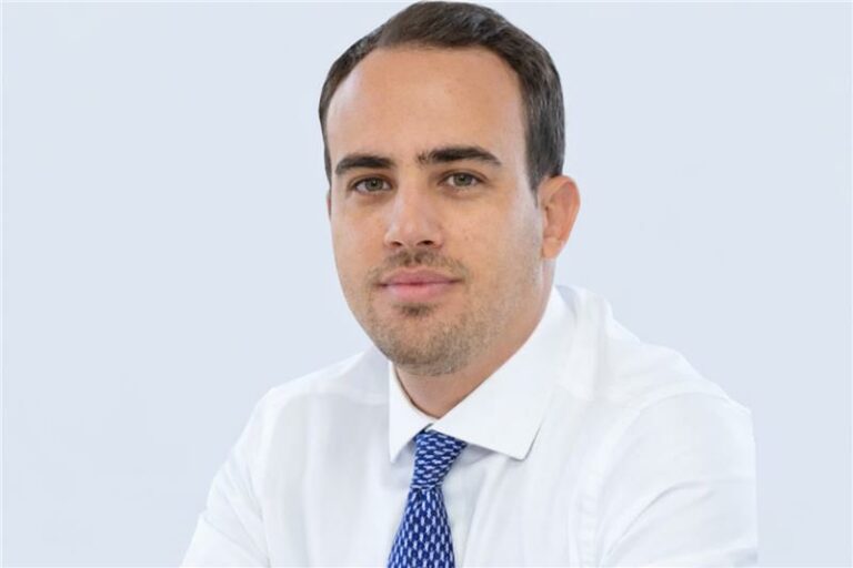 Pessoa vestindo uma camisa social branca e uma gravata azul com padrão discreto, sentada diante de um fundo claro e uniforme. A pose é formal, sugerindo um retrato profissional ou corporativo. (telemedicina)