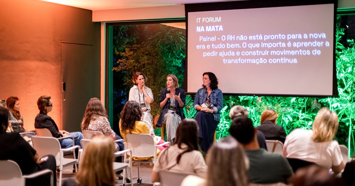 Um grupo de pessoas está reunido em uma sala para um painel de discussão. Três palestrantes sentam-se à frente do público, cada uma segurando um microfone enquanto conversam. Atrás delas, um telão exibe o título “IT Forum na Mata” e um texto sobre transformação contínua no RH. A sala tem grandes janelas que revelam vegetação iluminada do lado de fora. O público está sentado em fileiras de cadeiras brancas, atento ao debate.