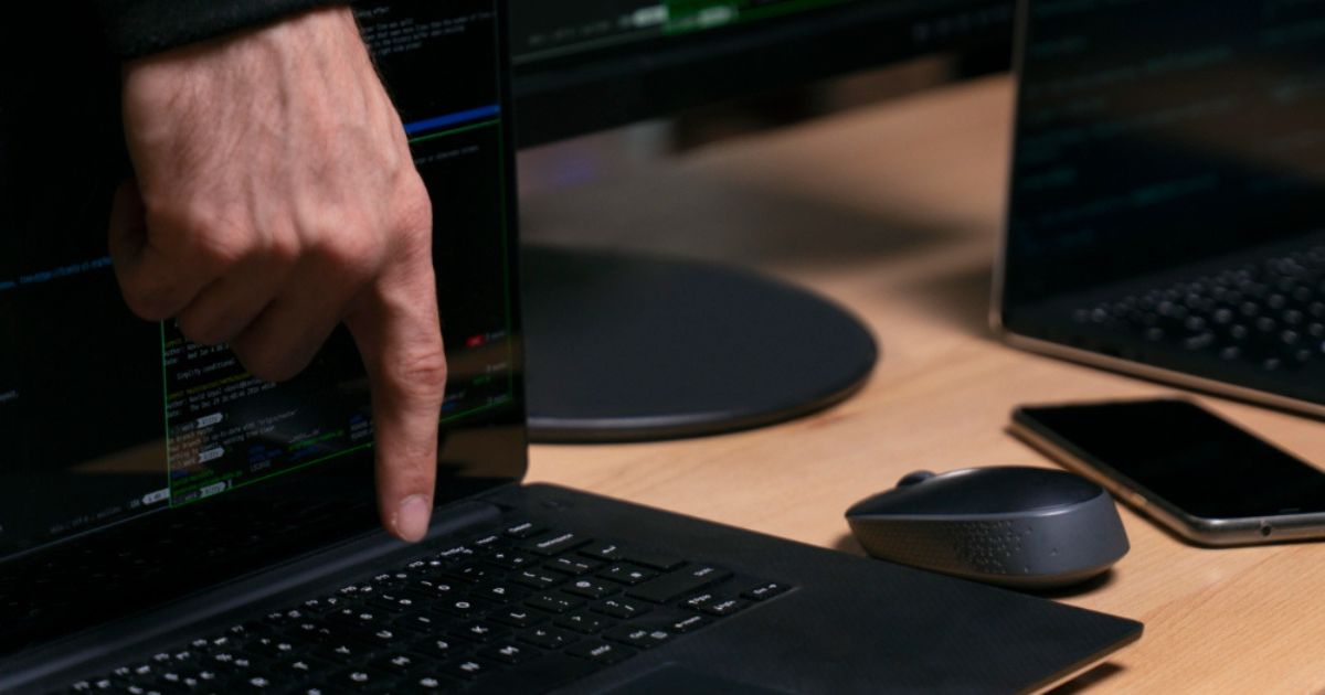 Uma mão aponta para o teclado de um laptop enquanto várias linhas de código são exibidas na tela. O computador está sobre uma mesa de madeira, ao lado de um mouse cinza, um smartphone e outro laptop parcialmente visível. Ao fundo, há monitores adicionais mostrando interfaces de programação, criando um ambiente típico de trabalho com desenvolvimento de software ou análise técnica. (Caso Epstein)