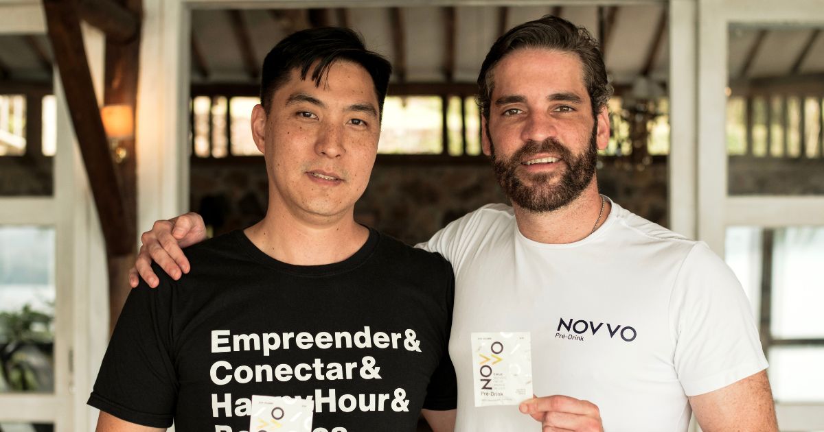 Duas pessoas posam lado a lado em um ambiente interno iluminado, com estruturas de madeira e vidro ao fundo. A pessoa à esquerda usa uma camiseta preta com texto branco em estilo de lista: “Empreender& Conectar& Happy Hour& Pessoas”. A pessoa à direita usa uma camiseta branca com o logo “NOVVO Pré-Obra” no peito. Ambas seguram pequenos cartões ou embalagens da marca “O2N” e uma delas apoia a mão no ombro da outra em gesto de amizade ou parceria.