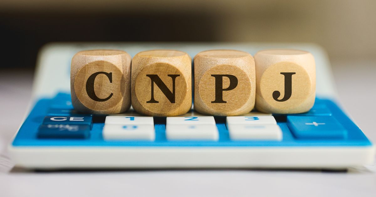Quatro blocos de madeira com letras impressas formando a sigla “CNPJ” estão posicionados sobre uma calculadora azul e branca, sugerindo uma temática relacionada a negócios, contabilidade ou cadastro empresarial.