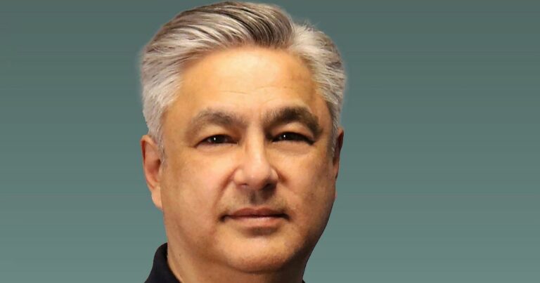 Pessoa com cabelo grisalho penteado para trás, vestindo uma camisa escura, posicionada diante de um fundo liso em tom verde-acinzentado. A composição é centrada na parte superior do corpo, com iluminação suave. (Inmetrics)