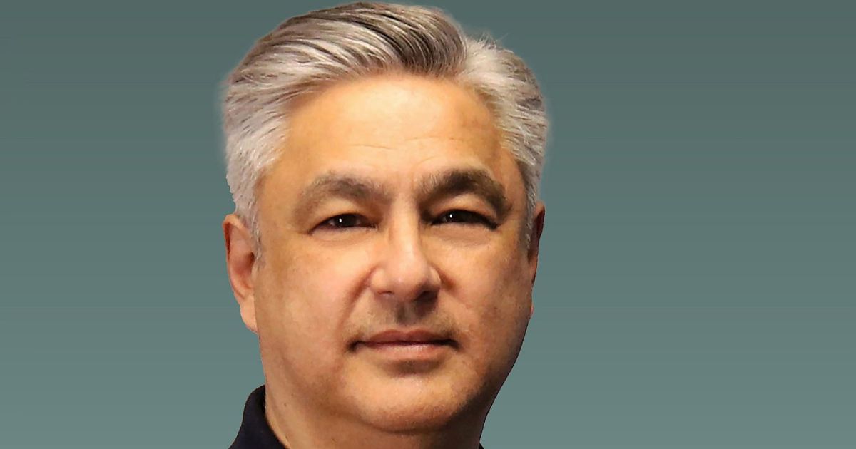 Pessoa com cabelo grisalho penteado para trás, vestindo uma camisa escura, posicionada diante de um fundo liso em tom verde-acinzentado. A composição é centrada na parte superior do corpo, com iluminação suave. (Inmetrics)