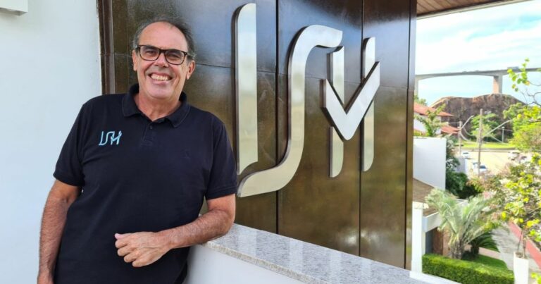 Pessoa usando uma camisa polo preta com um logotipo bordado no peito, apoiada em uma mureta de mármore claro. Ao fundo, há um grande painel metálico com o monograma “ISH” em destaque, além de uma área externa com árvores, jardins e estrutura arquitetônica visível à distância.