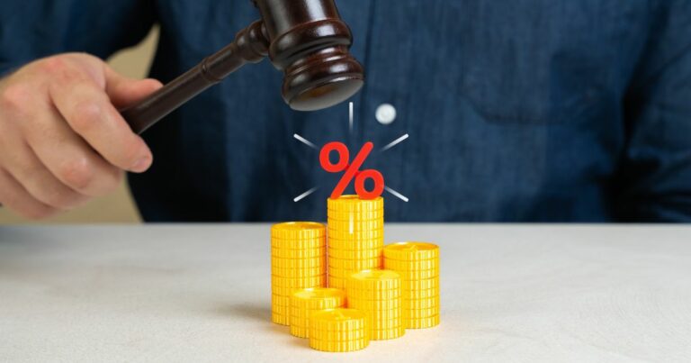 Uma mão segura um martelo de juiz prestes a bater sobre uma pilha de moedas douradas organizadas em diferentes alturas. Acima das moedas, há um símbolo vermelho de porcentagem, sugerindo temas relacionados a decisões judiciais, impostos, finanças ou regulamentação econômica. O fundo é simples, e a pessoa está vestindo uma camisa azul‑escura. (reforma tributária)